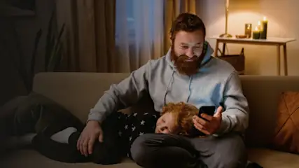 Ein Mann im grauen Hoodie sitzt auf einem Sofa und hält ein Smartphone; ein Kind liegt auf seinem Schoß. Im Hintergrund sind Kerzen.