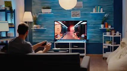 Ein Mann sitzt auf einem Sofa und spielt ein Videospiel auf einem Fernseher in einem Wohnzimmer.