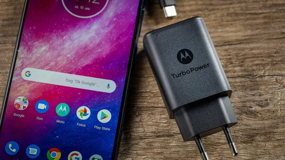Ein Smartphone mit farbigem Hintergrund und App-Symbolen liegt neben einem schwarzen Motorola TurboPower-Ladegerät auf Holz.