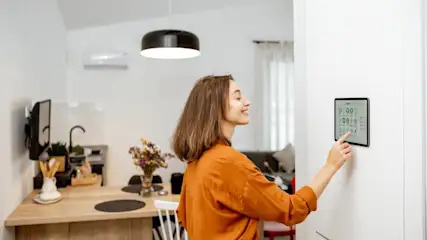 Eine Frau tippt auf ein digitales Display an einer weißen Wand in einem hellen Raum mit Tisch und Lampe.