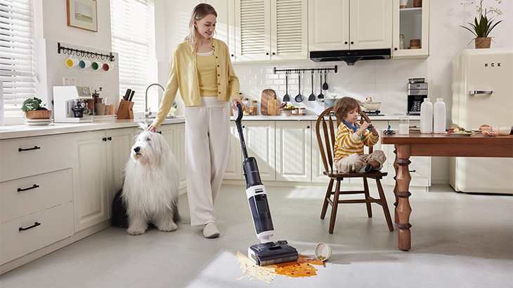 Frau reinigt mit Staubsauger Küche mit Esstisch, Barhockern, Fenster und Katzenspielplatz. Flüssigkeit auf grauem Boden.