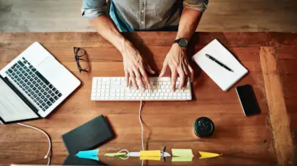 Draufsicht auf eine Person, die an einem Schreibtisch mit Laptop, Notizbuch, Stift, Handy, Brille, Notizen und Kaffee tippt.
