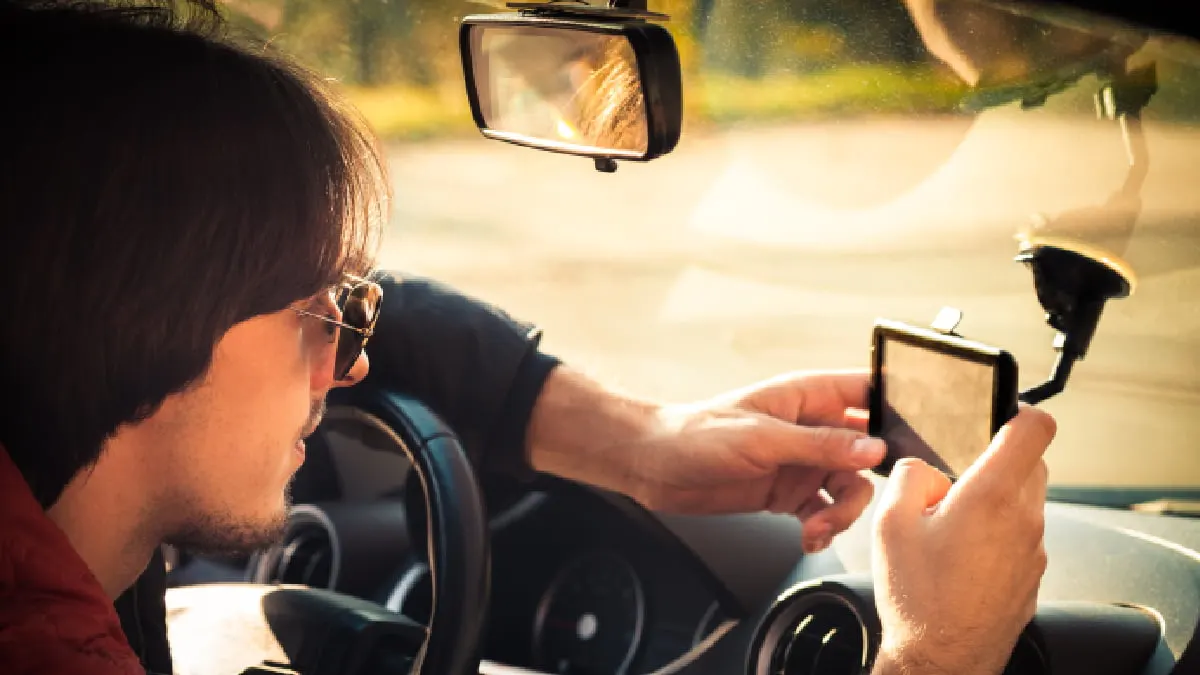 Ein Mann mit Sonnenbrille sitzt im Auto, hält ein GPS-Gerät und blickt auf die Straße. Der Rückspiegel ist sichtbar.