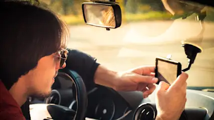 Ein Mann mit Sonnenbrille sitzt im Auto, hält ein GPS-Gerät und blickt auf die Straße. Der Rückspiegel ist sichtbar.