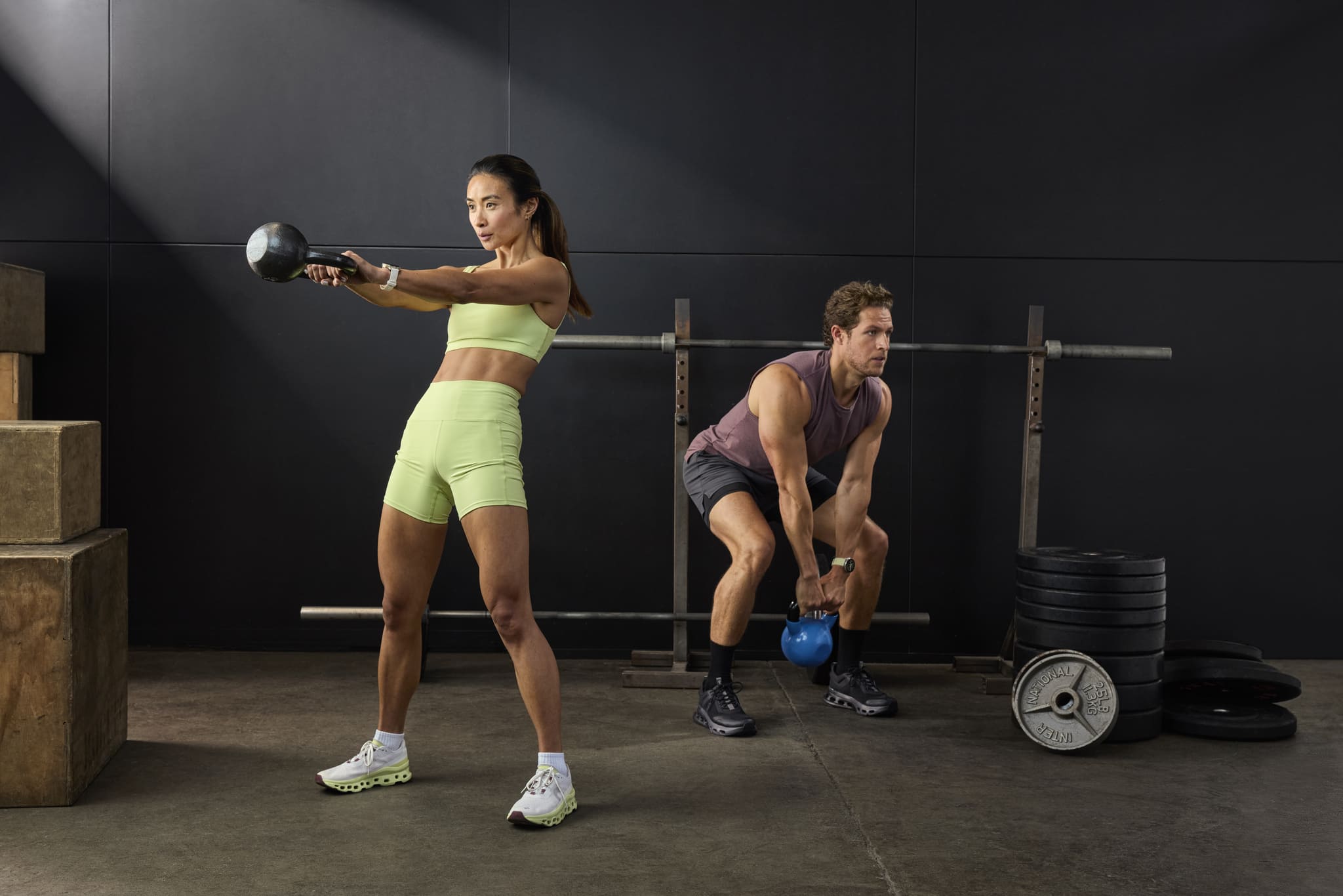 Zwei Personen trainieren im Fitnessstudio mit Kettlebells, umgeben von Hantelscheiben und Trainingsboxen.