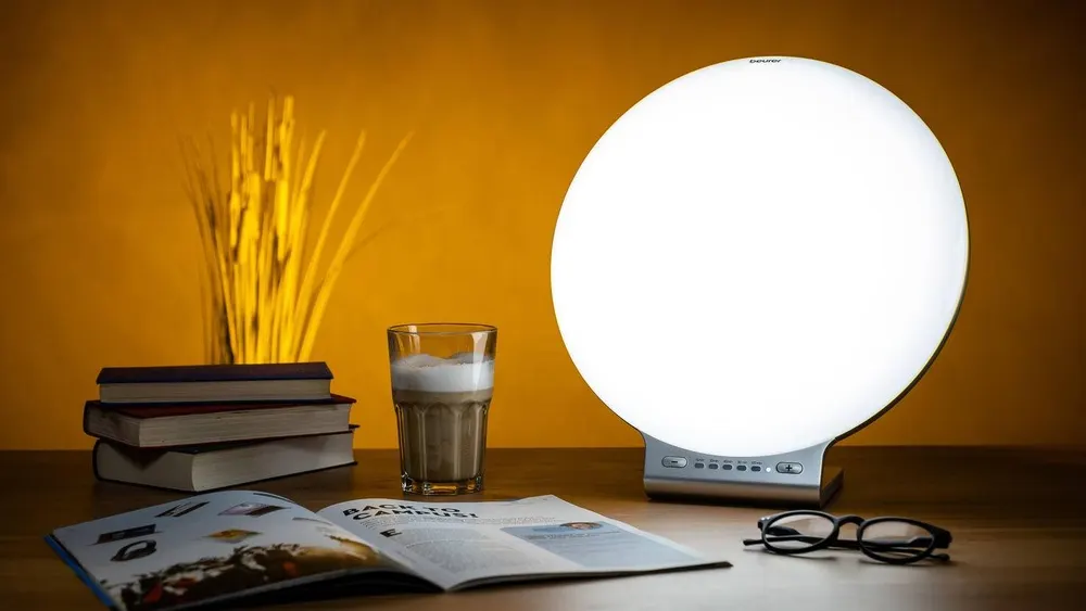 Tisch mit Lampe, Büchern, Zeitschrift, Getränk und Brille vor ockerfarbenem Hintergrund.