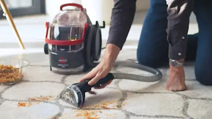 Eine Person reinigt einen Teppich mit einem tragbaren Teppichreiniger. Auf dem Teppich sind Flecken. Eine Glasschale steht daneben.