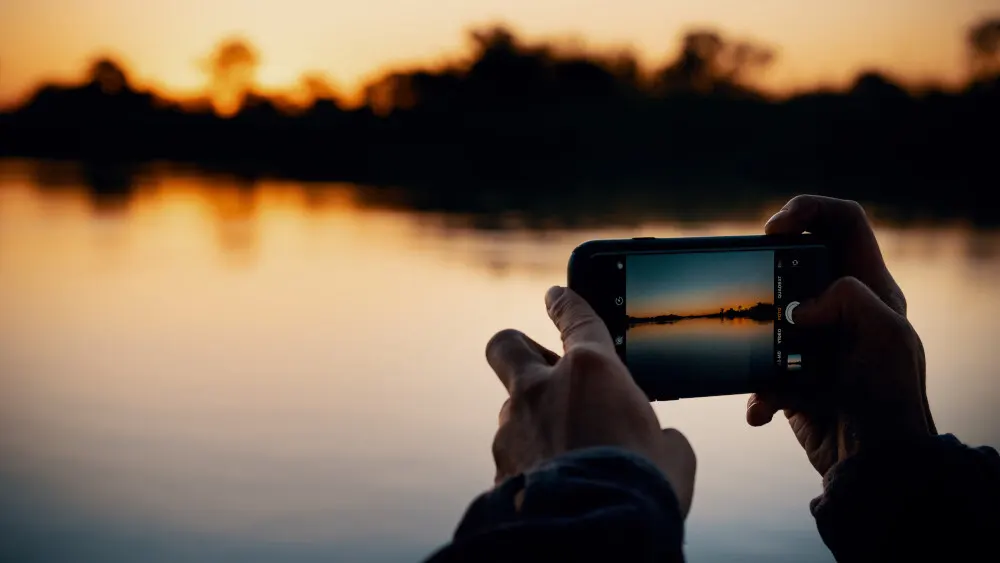 Hände halten ein Smartphone, das einen See bei Sonnenuntergang mit Bäumen im Hintergrund zeigt. Das Wasser reflektiert den Himmel.