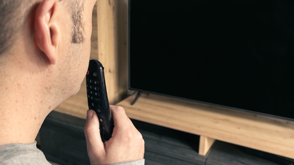 Ein Mann spricht in eine Fernbedienung und blickt auf einen Fernseher.