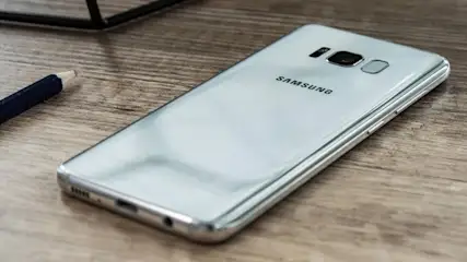 Silbernes Samsung-Smartphone liegt auf einem Holztisch. Eine Bleistiftspitze und ein Teil eines schwarzen Objekts sind auch sichtbar.