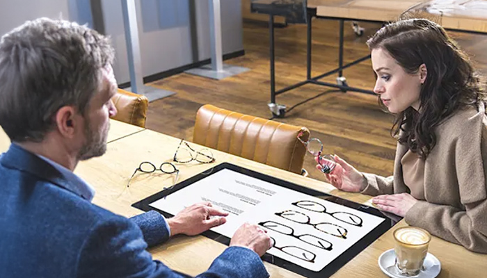Ein Mann und eine Frau sitzen an einem Tisch und betrachten Brillenmodelle auf einem Blatt Papier. Eine Tasse Kaffee steht daneben.