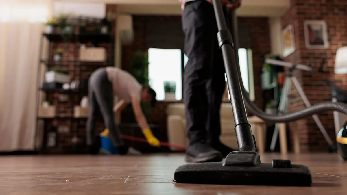 Jemand saugt mit einem Staubsauger den Holzboden im Wohnzimmer. Im Hintergrund putzt eine zweite Person.