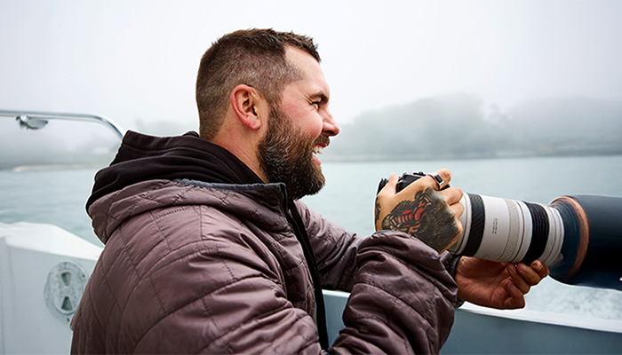 Fotograf mit Teleobjektiv am nebligen See; seine tätowierte Hand hält die Kamera, gesteppte Jacke, Wasser und Bäume im Hintergrund.