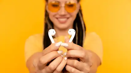 Frau mit gelber Brille hält zwei weiße Ohrhörer vor einem gelben Hintergrund.
