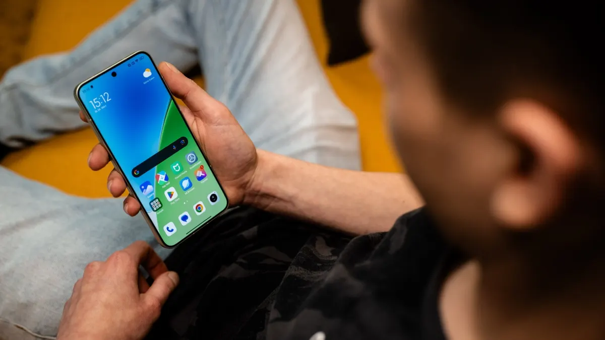 Eine Person hält ein Smartphone mit blau-grünem Bildschirm in der Hand, während sie auf einer gelben Oberfläche sitzt.