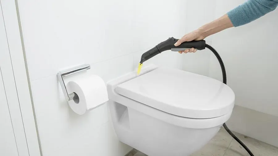 Eine Person reinigt eine weiße Toilette mit einem schwarzen Dampfreiniger, gelber Dampf ist sichtbar. Eine Toilettenpapierrolle hängt an der Wand.