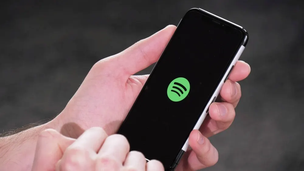 Eine Hand hält ein Smartphone mit schwarzem Bildschirm und grünem Spotify-Logo vor einem dunklen Hintergrund.