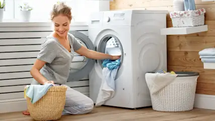 Eine Frau kniet vor einer weißen Waschmaschine und räumt blaue Wäsche in einen Korb. Im Hintergrund sind Regale und Körbe.