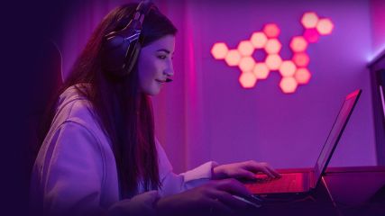 Eine junge Frau ist seitlich zu sehen. Sie sitzt an einem Laptop, hat ein Headset an und ihre Hände liegen auf Maus und Tastatur. Die Hintergrundbeleuchtung ist lila.