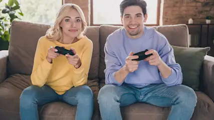 Eine Frau in Gelb und ein Mann in Lila sitzen auf einem Sofa und halten schwarze Gamecontroller.