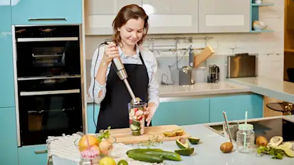 In einer Küche mixt eine Frau mit einem Stabmixer Zutaten in einem Glas. Verschiedene Früchte und Gemüse liegen auf der Theke.