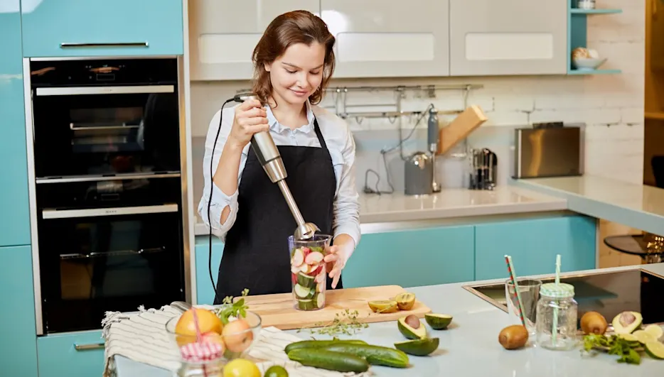 In einer Küche mixt eine Frau mit einem Stabmixer Zutaten in einem Glas. Verschiedene Früchte und Gemüse liegen auf der Theke.