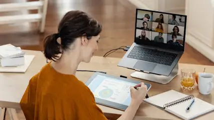 Eine Person arbeitet an einem Tisch mit Laptop, Grafiktablett, Notizbuch und Getränken während eines Videoanrufs.
