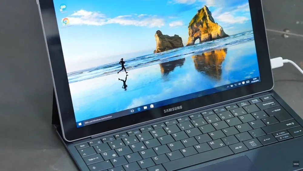 Ein Samsung-Tablet mit Tastatur zeigt ein Strandbild mit einer Person, die am Ufer entlangläuft, unter blauem Himmel. Ein Ladekabel ist angeschlossen.