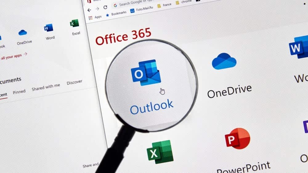 Eine Lupe vergrößert das Outlook-Logo am PC-Monitor.