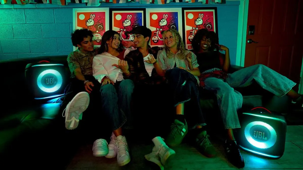 Fünf junge Menschen sitzen auf einem Sofa zwischen zwei Modellen der JBL PartyBox Encore 2.