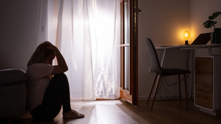 Eine Person sitzt auf dem Boden neben einem Sofa, mit einem Schreibtisch, einem Stuhl und einer Lampe im Hintergrund.