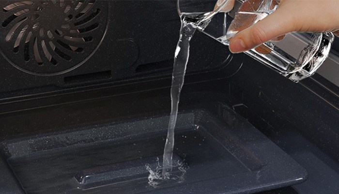 Eine Hand gießt Wasser aus einem Glas in einen schwarzen Ofen mit einem Ventilator an der Oberseite.