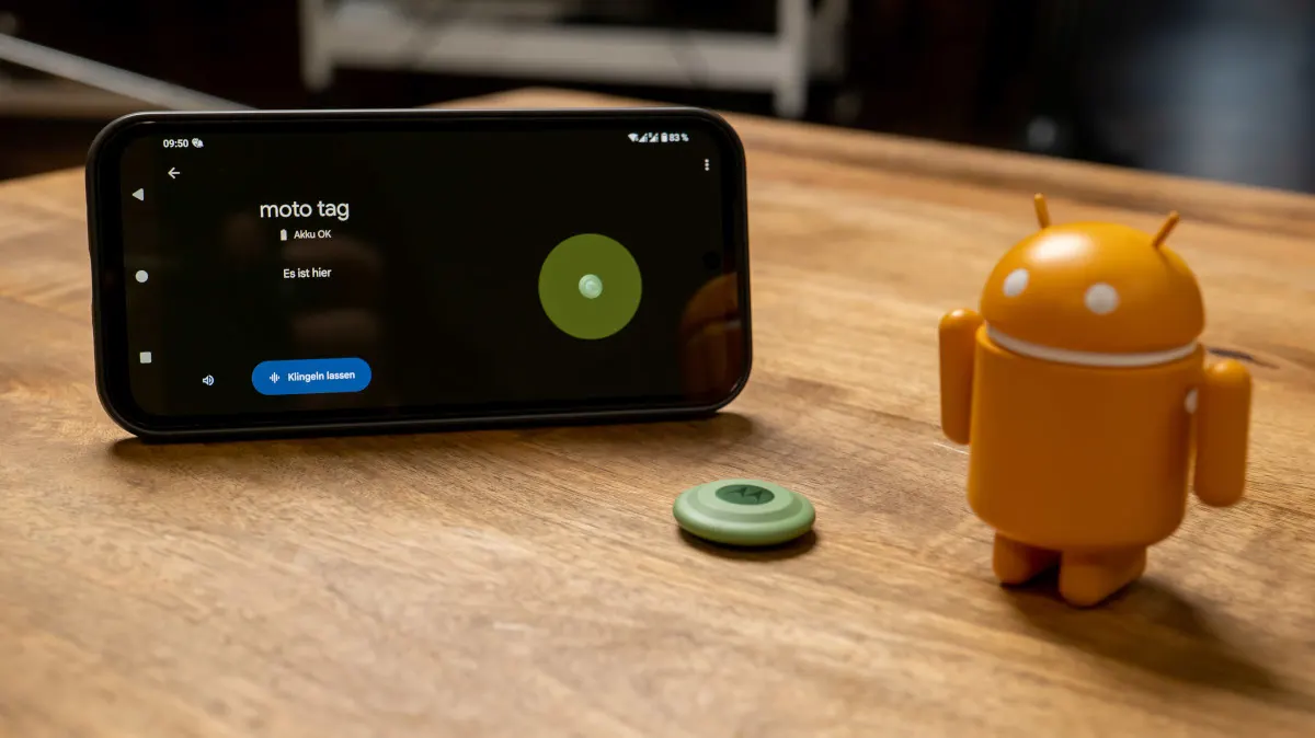 Ein Smartphone mit der Aufschrift "moto tag", ein grüner Tracker und eine orangefarbene Android-Figur auf einem Holztisch.