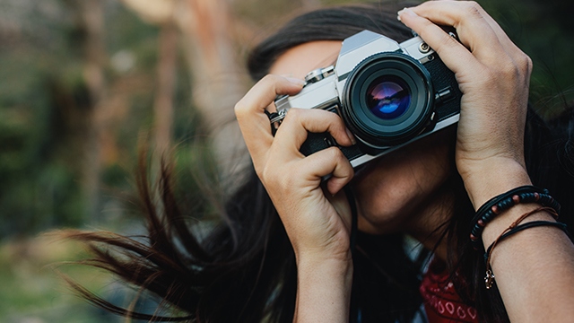 Eine Person hält eine silberne und schwarze Kamera vor ihr Gesicht, im Hintergrund ist eine grüne Vegetation zu sehen.