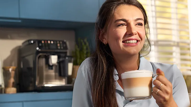 Frau lächelt und hält einen Latte Macchiato in einem Glas. Im Hintergrund sind Küchengeräte sichtbar.