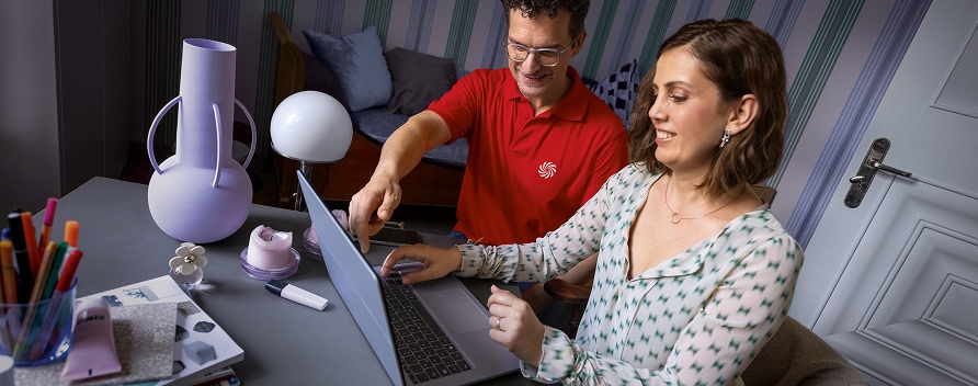Mitarbeiter im roten Poloshirt erklärt einer Kundin lächelnd die Bedienung eines Tablets zu Hause.