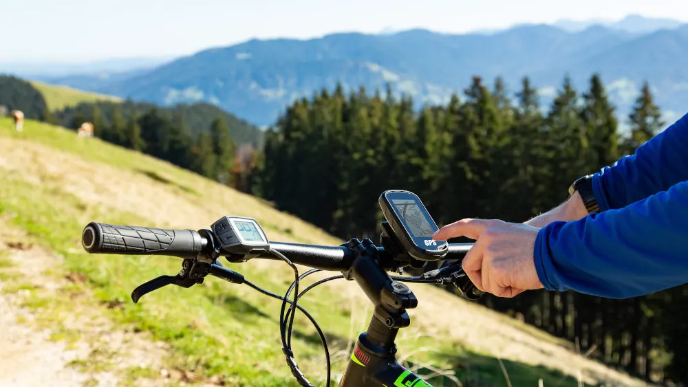 Fahrradlenker mit GPS-Gerät vor Hügeln und Bäumen. Eine Hand bedient das GPS. Kühe grasen im Hintergrund auf der Wiese.