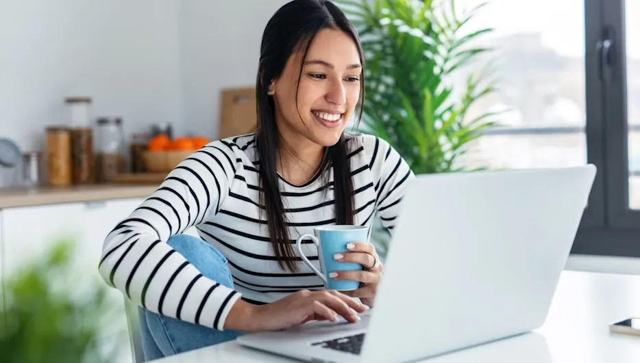 Frau mit gestreiftem Oberteil und Jeans sitzt am Tisch und benutzt Laptop. Sie hält eine blaue Tasse, Zimmerpflanze im Hintergrund.