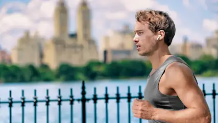Ein Mann mit grauem Tanktop und Kopfhörer joggt vor einer schwarzen Gitterzaun und einem See mit Gebäuden im Hintergrund.