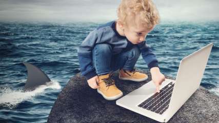 Ein Junge sitzt mit einem Laptop auf einem Felsen im Meer und wird von einem Hai umkreist.