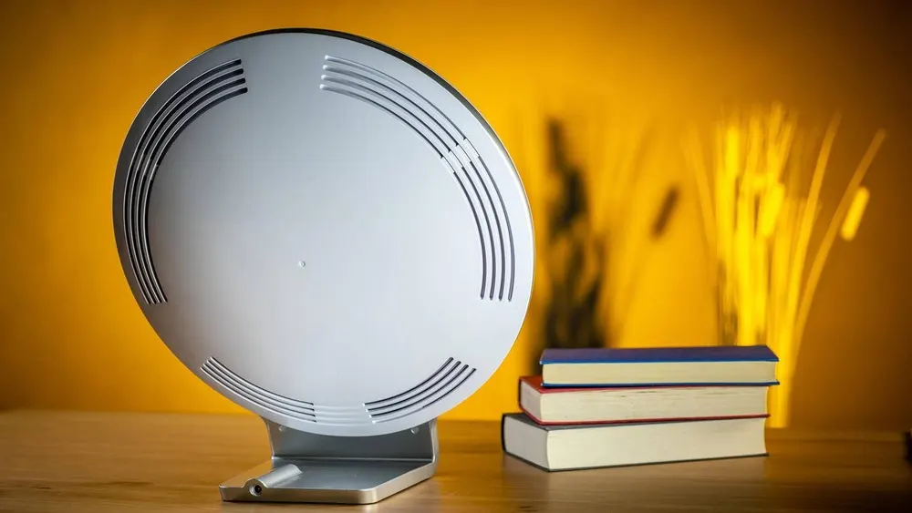 Runder, silberne Tageslichtlampe auf einem Tisch, daneben ein Stapel Bücher und dekorative Äste vor gelbem Hintergrund.