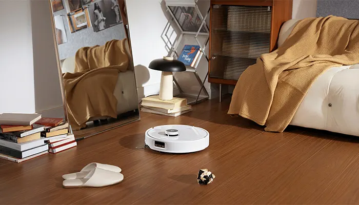 Ein weißer Roboterstaubsauger auf einem Holzboden in einem Wohnzimmer mit Büchern, Pantoffeln und Möbeln.