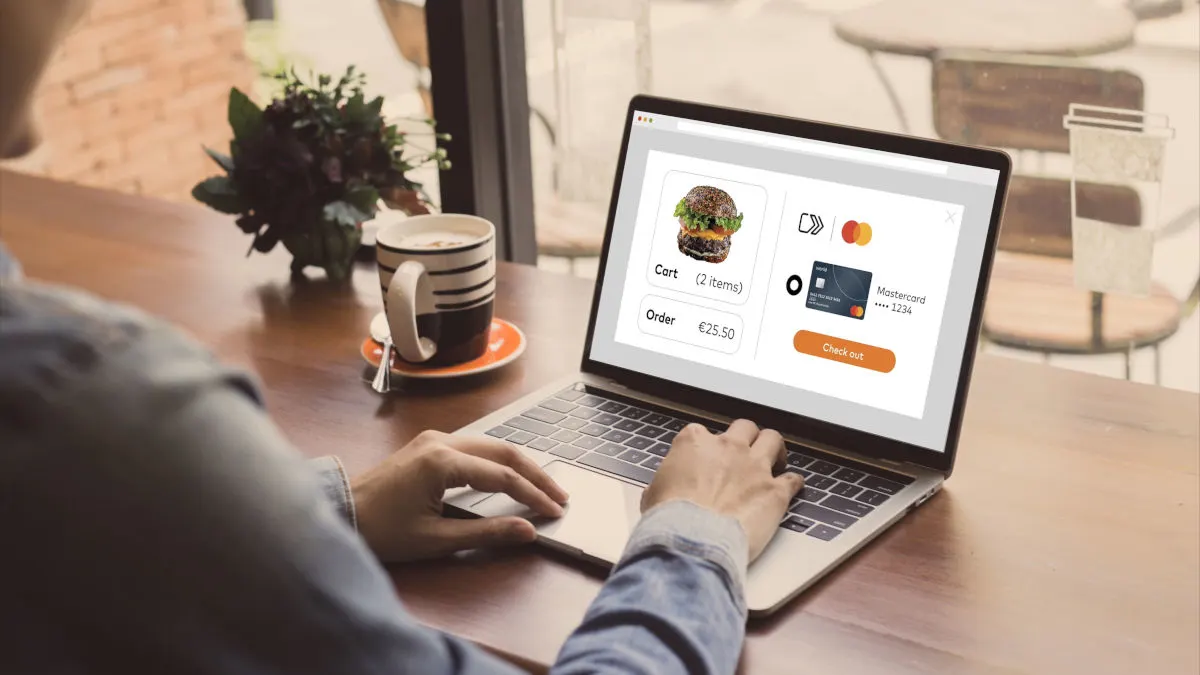Eine Person nutzt einen Laptop für eine Online-Bestellung mit einem Burger und einer Mastercard auf dem Bildschirm. Daneben stehen Kaffee und Blumen.