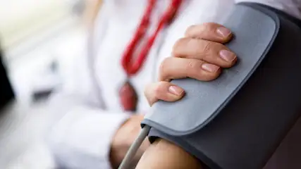 Eine Hand hält ein graues Blutdruckmessgerät am Arm, mit einer roten Kordel und einem weißen Kittel im Hintergrund.