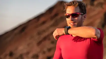 Ein Mann mit rotem Oberteil und Armbanduhr steht vor einem braunen Felsen und trägt eine rote Sonnenbrille.