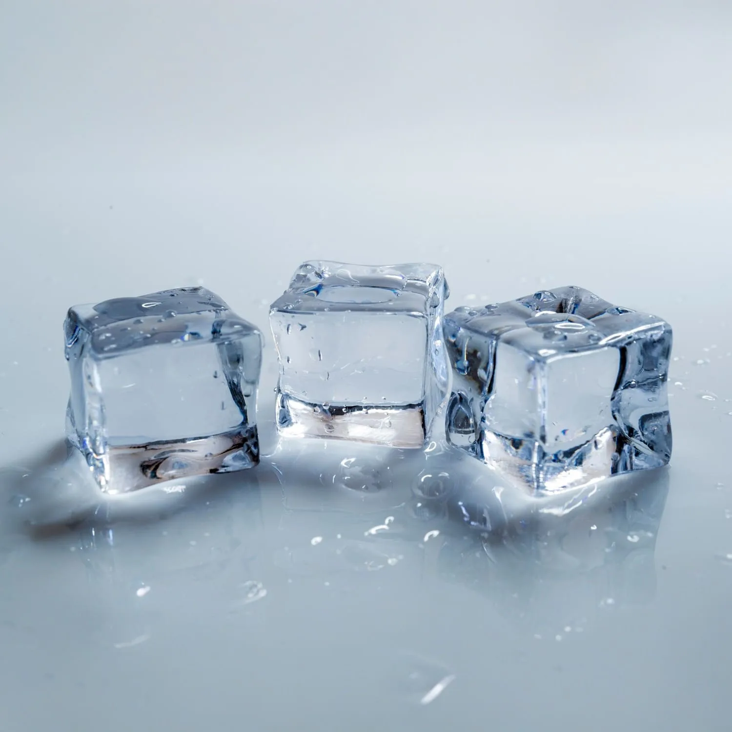 Drei klare Eiswürfel liegen auf einer hellen Oberfläche mit kleinen Wassertropfen um sie herum.
