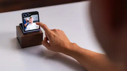 Eine Hand berührt einen kleinen Bildschirm auf einem Holzsockel. Auf dem Bildschirm ist ein Mann zu sehen, der von einer anderen Person fotografiert wird.