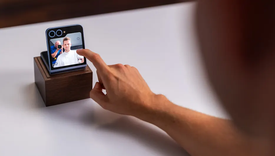 Eine Hand berührt einen kleinen Bildschirm auf einem Holzsockel. Auf dem Bildschirm ist ein Mann zu sehen, der von einer anderen Person fotografiert wird.