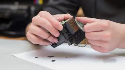 Eine Person reinigt einen schwarzen Druckkopf mit einem Tuch über einem weißen Blatt Papier mit Tintenflecken.