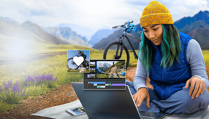 Frau mit gelber Mütze arbeitet an einem Laptop im Freien. Fahrrad und Berge im Hintergrund. Videobearbeitungssoftware auf dem Bildschirm.